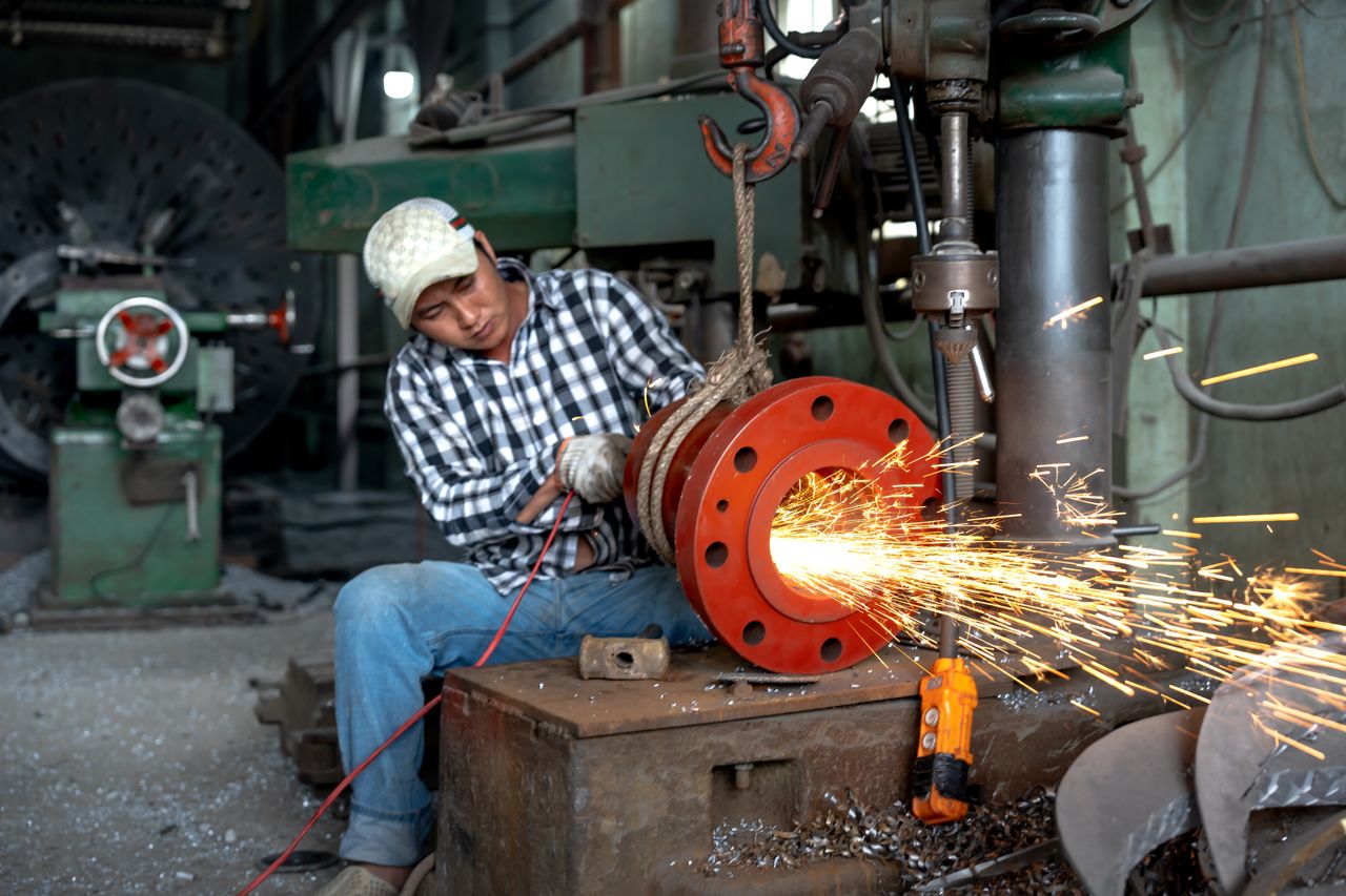 about-us-img An Asian blue-collar worker operates machinery, grinding metal with sparks flying in a warehouse setting.